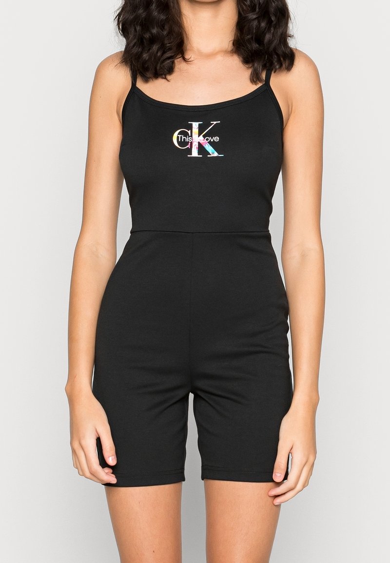 Femme portant une combinaison noire sans manches avec de fines bretelles et un logo multicolore « CK This Love » sur la poitrine.