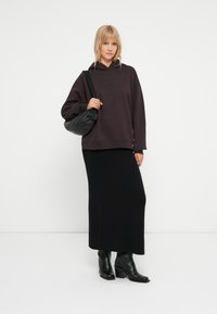 Tmavošedá oversize mikina s rebrovanými manžetami, v kombinácii s čiernou pletenou maxi sukňou. Modelka drží malú čiernu tašku a má obuté čierne ihlové čižmy.