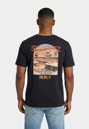 Homme portant un T-shirt noir avec un graphisme au dos montrant deux personnes en train de bronzer sur un bateau dans un port, texte "ST TROPEZ" et "HERO7".