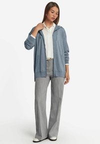 Ljusblå huvtröja i mjukt material, med långa ärmar och framfickor, bärs över en vit skjorta och grå vida byxor.