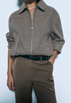 Veste marron à fermeture éclair avec col, en tissu texturé, associée à un pantalon marron lisse et une ceinture noire.