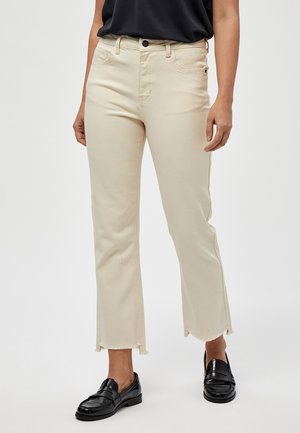 Beige straight-leg jeans with raw hem, mid-rise waist, five pockets, and a button fly. Worn with black leather loafers.
