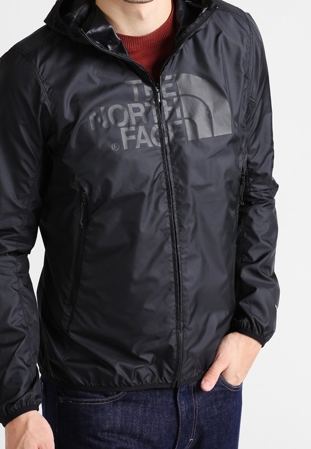 Homme portant une veste zippée noire The North Face avec capuche sur un pull rouge, avec une main dans la poche d'un jean bleu.