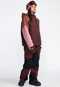 Snowboard-outfit med en jakke og bukser i farveblokke af vinrød og pink, med tekstureret stof og tætsiddende design, suppleret med goggles og en hue.