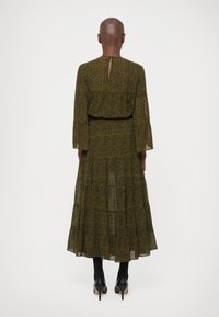 Robe longue à étages de couleur vert foncé avec un motif floral jaune. Dispose de manches longues, d'un dos en clé et d'un tissu fluide et léger. Escarpins noirs.