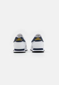 Baskets blanches avec talon et accents bleu marine, dotées d'une tige texturée, d'un bout arrondi et d'un logo "new balance" jaune vif à l'arrière.