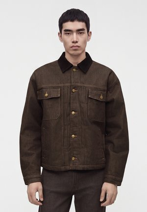 Joven con chaqueta de mezclilla marrón con cuello negro y botones dorados, de pie frente a un fondo liso y claro.