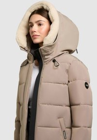 Frau trägt eine beige gesteppte Jacke mit einer großen, mit Fleece gefütterten Kapuze und Reißverschluss, leicht nach links gewandt vor einfachem Hintergrund.