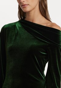 Πράσινο βελούδινο φόρεμα με σχέδιο που αφήνει τους ώμους ακάλυπτους, διαθέτοντας απαλό ύφασμα και κρεμαστή λαιμόκοψη. Οι μακριές μανίκες τονίζουν τη κομψή σιλουέτα.