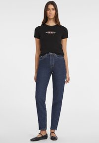 Čierne tričko s logom "GUESS JEANS", kombinované s vysokými tmavomodrými džínami, ktoré majú klasický strih a priame nohavice, nosené s čiernymi plochými topánkami.
