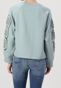 Sweat-shirt bleu clair à manches longues avec broderies florales et accents métalliques sur les épaules. Coupe douce et décontractée avec ourlet côtelé.