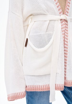 Biały dziergany kardigan z różową lamówką, dużą przednią kieszenią i paskiem związanym w talii, noszony z niebieskimi dżinsami.