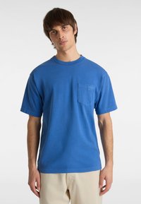 Camiseta de algodón azul con mangas cortas, escote redondo y un bolsillo en el pecho. Textura suave, corte casual, combinada con pantalones beige claros.