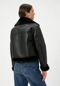 Giacca nera in pelle corta con colletto e polsini in pelliccia sintetica. Presenta una texture liscia e un design semplice, abbinata a jeans blu.