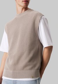 Gilet beige a maglia con scollatura e orlo a coste, caratterizzato da un motivo testurizzato. Indossato sopra una t-shirt bianca, con un piccolo logo dettagliato sull'orlo.