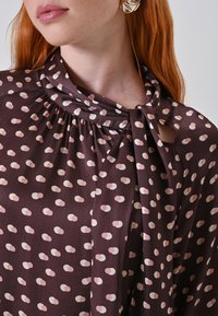 Blusa marrone con pois rosa chiaro, caratterizzata da una scollatura arricciata e un dettaglio annodato sul davanti. Tessuto liscio con una leggera lucentezza.