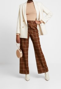 Blazer beige, top a coste color sabbia, pantaloni alti a quadri marroni, stivaletti crema alla caviglia e borsa tonda intrecciata con accenti metallici.
