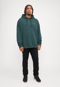 Sudadera verde con capucha, con un bolsillo frontal y un pequeño logo dorado, combinada con pantalones negros y zapatillas oscuras. Textura suave, corte relajado.