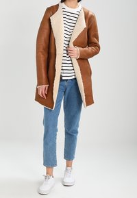 Abrigo de cuero marrón con forro de borrego crema, diseño de apertura frontal, mangas largas, combinado con una camisa a rayas y jeans azules. Zapatillas blancas.