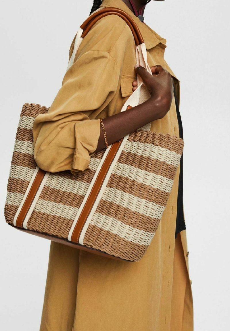 Personne en manteau beige portant un grand sac cabas tissé beige et marron rayé avec des poignées en cuir sur l'épaule.