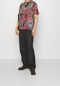 Man med en röd och svart leopardmönstrad t-shirt, svarta byxor och mörkbruna dressskor, som håller en svart mapp.