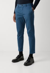 Kék chino nadrág, taperd szabású, pamutból készült. Gombos záródással, oldalsó zsebekkel rendelkezik, és fekete fűzős cipővel van párosítva.