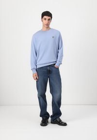 Sudadera azul claro con cuello redondo y pequeño logo bordado; combinada con jeans azul oscuro y zapatos negros, de pie contra un fondo simple.
