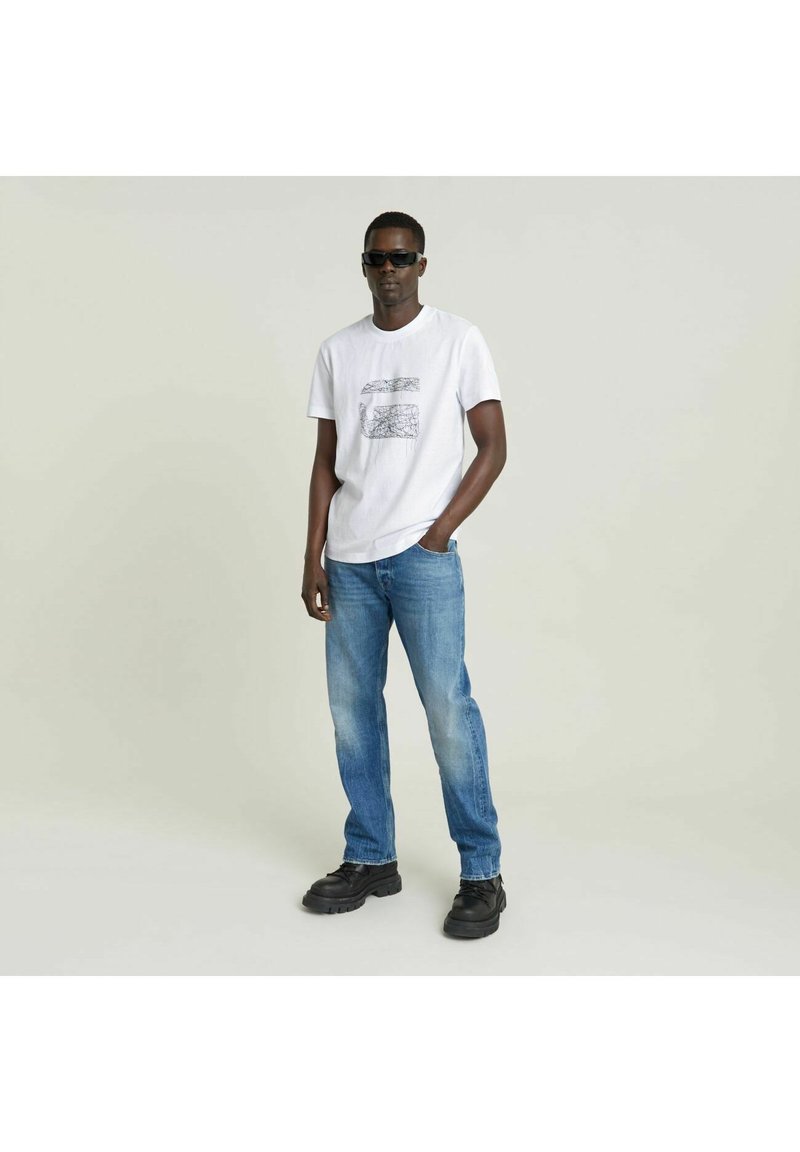 Camiseta blanca de algodón con un diseño gráfico negro, combinada con jeans de mezclilla azul claro y zapatos negros de suela gruesa. Unas gafas de sol negras completan el look.