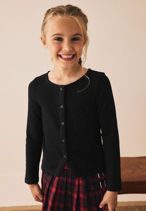 Schwarzes langärmliges, geripptes Strickjacke mit Rundhalsausschnitt und vorne mit Knöpfen zu schließen, kombiniert mit einem roten karierten Rock mit verstellbaren Trägern.