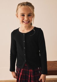 Cardigan noir à manches longues en maille côtelée avec encolure ronde et fermeture à boutons sur le devant, associé à une jupe rouge à carreaux avec bretelles réglables.