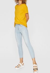 Camiseta de algodón amarilla, jeans ajustados de mezclilla de color azul claro con detalles rasgados, y sandalias blancas de tiras con acentos plateados.