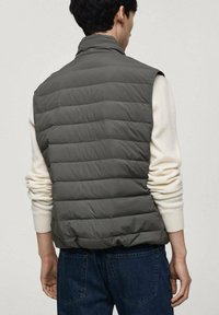 Grå pufferväst med kviltad textur, hög krage och ärmlös design, bärs över en krämfärgad långärmad skjorta och blå jeans.