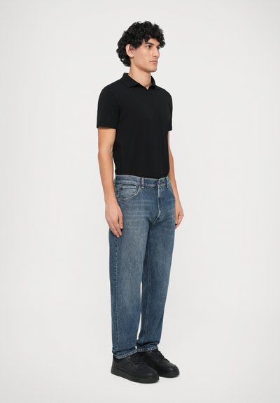 Polo shirt nero a maniche corte, jeans in denim blu e sneakers nere. Fit dritto con dettagli minimi, indossato in una posa in piedi.