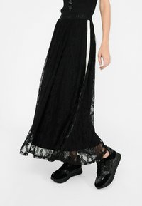 Schwarzer Spitzen-Maxikilt mit floralem Muster, transparenter Überlage und weißem Seitenstreifen; kombiniert mit glänzenden schwarzen Plateau-Sneakern.