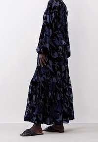 Robe maxi à manches longues en bleu foncé avec un motif floral, dotée d'un ourlet à volants et d'un tissu texturé. Associée à des sandales à glissière noires.