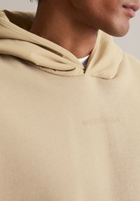 Beżowa bluza z kapturem o teksturowanym wykończeniu i luźnym kroju. Posiada kaptur oraz subtelne logo w tkaninie. Miękki, gładki materiał.