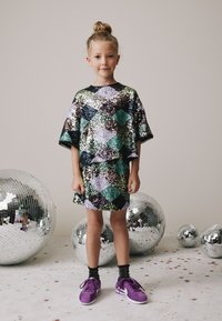 Paljettklänning i diamantmönster med gröna, lila och svarta färger. Matchas med lila sneakers. Reflekterande discokulor i bakgrunden.