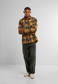 Gul og svart rutete flanellskjorte med knappedetaljer, kombinert med mørke denimbukser og lyse sneakers, vist mot en nøytral bakgrunn.