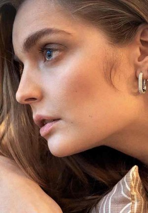 Profilsyn af en person med langt brunt hår, lys hud, iført hoop øreringe og bløde lyserøde læber. Baggrunden har et struktureret mønster.