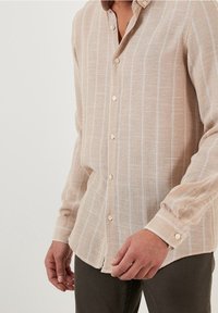 Světlá béžová košile na knoflíky s bílými svislými pruhy, dlouhými rukávy a měkkou texturou. Má zapínání na knoflíky a manžety.