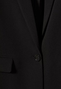 Gros plan d'un blazer noir montrant un seul bouton noir et une poche à rabat sur le côté gauche.