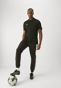 Μαύρο πόλο πουκάμισο με λευκό λογότυπο, μαύρο παντελόνι φόρμας με λευκά πλαϊνά στοιχεία και μαύρα ποδοσφαιρικά παπούτσια, στέκεται πάνω σε μια ποδοσφαιρική μπάλα.