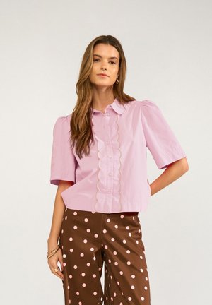 Junge Frau mit langen Haaren trägt eine hellrosa Bluse mit gewellter Front und braune Hose mit hellrosanen Punkten.