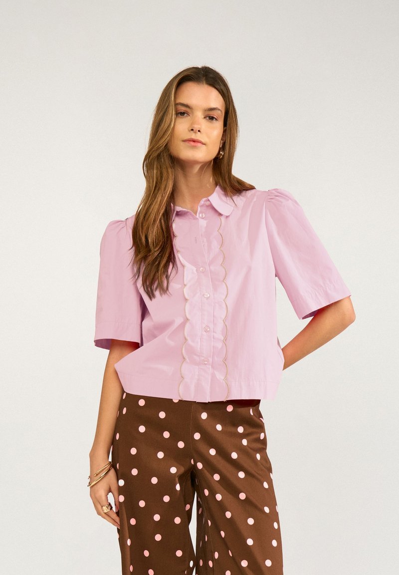 Jeune femme aux longs cheveux portant un chemisier rose clair avec un détail festonné sur le devant et un pantalon marron à pois rose clair.