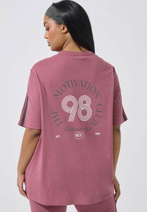 Frau mit langen dunklen Haaren, die ein lockeres mauvefarbenes T-Shirt trägt, auf dessen Rückseite "THE MOTIVATION CLUB 98" in weißem und schwarzem Text gedruckt ist.