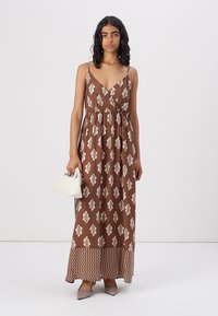 Robe maxi marron avec motif de feuilles blanches, décolleté en V, fines bretelles, taille froncée et ourlet texturé avec un design en zigzag. Le modèle tient un sac à main blanc.