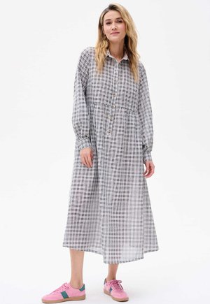 Langes, knopfbares Kleid mit einem schwarz-weißen Karomuster. Verfügt über lange Ärmel und eine geraffte Taille. Kombiniert mit pinken Turnschuhen.