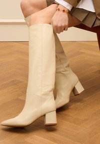 Beige leren kniehoge laarzen met een spitse neus en blokhak. De oppervlakte is glad, wat de naden en het aansluitende ontwerp benadrukt.