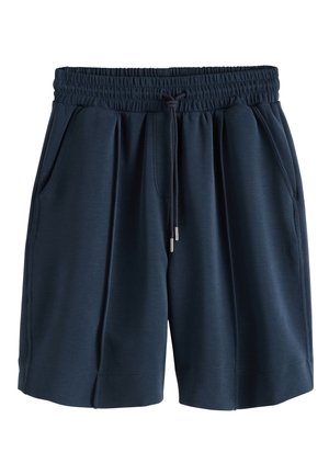 Femme portant un haut en maille beige à manches courtes et un short bleu marine à cordon, tenant un sac à main macramé beige avec des anses marron.