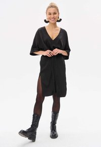 Schwarzes Satin-Kleid mit tiefem V-Ausschnitt, Fledermausärmeln und einem Bindegürtel; kombiniert mit blickdichten Strumpfhosen und klobigen schwarzen Schnürstiefeln.